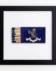 A framed display showcases a vintage matchbox with a dark blue cover, featuring a gold lion holding a globe. Half the matches are visible, with blue tips. The matchbox banner reads, "The Ritz London." The black frame from Match South's Art Square Blk Frame collection, crafted from clear acrylic for modern elegance, is placed against a white background.