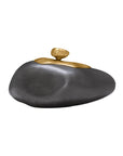 The Import Collection’s Finkle Large Box is a decorative piece resembling a smooth, dark gray stone crafted from aluminum, topped with a gold pebble-shaped accent and set against a white background.