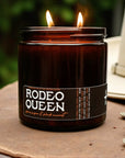 A brown glass Faire candle labeled "Black Currant & Spun Sugar" with two flames burns outdoors. A white object, possibly a hat, is partially visible in the background.