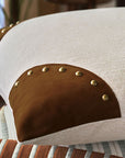 A close-up of the Eastern Accents RUFUS CORNER PILLOW shows leather corner details with bold brushed gold nailheads. The beige pillow sits atop a patterned cushion with white, blue, and brown stripes.