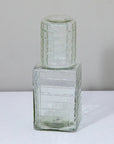 The Be Home Ruffle Glass Bedside Set includes a clear, square carafe with a matching lid, both with a grid pattern. Stylish and chic, it's the perfect nightstand accessory, shown here on a white surface against a light gray background.