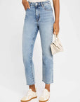 A person wears FRAME’s The Ruler Crop high-waisted, slim-straight jeans in light blue, paired with white sneakers with beige accents and a small beige handbag. The photo focuses on their lower half against a plain background.