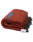The Faire Rust Red Baja Thunderbird Mexican Blanket features a rust hue with black fringe, a small blue design, and a white "west path WO" tag. Handmade in Mexico.