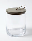 The BB-6.60026 Rustic Canister MD by Global Views features clear glass, a round flat beige lid, and a looped brown leather handle—ideal for modern or rustic kitchen storage—displayed against a white background.