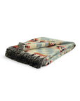 A folded Faire Secoya Throw with fringed edges, displaying a geometric pattern in muted green, beige, blue, and red.