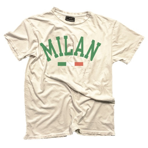 A wrinkled beige MILAN TEE with the word &quot;MILAN&quot; printed in green letters across the chest, accompanied by a small Italian flag below the text, lying flat on a white background in a Scottsdale Arizona bungalow by Wildcat Retro Brands.