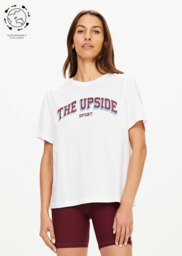 A woman models The Upside ALUMNI JODHI TEE in organic cotton with burgundy athletic shorts, standing before a white background. A “Sustainably Focused” icon is displayed in the top left corner.