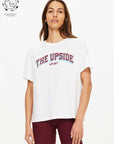 A woman models The Upside ALUMNI JODHI TEE in organic cotton with burgundy athletic shorts, standing before a white background. A “Sustainably Focused” icon is displayed in the top left corner.