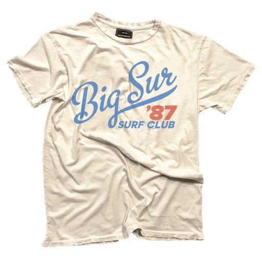 A crumpled light gray BIG SUR SURF CLUB TEE with &quot;Big Sur 987 Surf Club&quot; printed in blue and red on the front, displayed against a bungalow-style background.