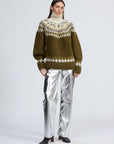 A woman with long brown hair and a neutral expression models the Rag & Bone beth oversized fair isle tneck sweater, featuring green and white patterns, paired with silver metallic pants and black shoes against a plain background.