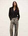 A woman with long, wavy dark hair stands against a neutral background, wearing the BA&SH Shirt Itsey— a black button-up with loose shoulders—tucked into high-waisted, loose grey jeans and styled with a silver studded belt. Hands in pockets.