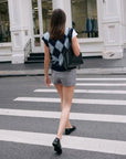 A woman with straight brown hair, in a White + Warren Mohair Luxe Argyle Vest, gray shorts, and black shoes, carries a black bag as she crosses a city street at a crosswalk.