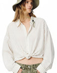 A person in the Denimist TWISTED FRONT SHIRT—a relaxed-fit linen-cotton top with thin vertical stripes and a front tie—paired with a wide-brimmed khaki hat and olive green pants stands against a white background, hands in pockets.