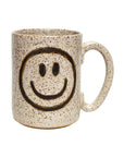 The Faire Ceramic Mug is a beige speckled handmade mug with a large handle and a black smiley face on the side. Dishwasher safe, it adds charm and practicality to any ivory mug collection.