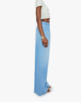 A person stands sideways against a white background, wearing Mother’s The Kitchen Drawer Skimp high-rise wide leg jeans in a recycled cotton blend with an elastic waistband, paired with a white cropped top, sandals, and a gold wristwatch.