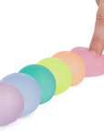 A hand squeezes a pink Mini Gump from Faire's 6-pack, with the pastel office fidgets lined up neatly on a white surface.