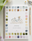 The Faire Spring watercolor workbook is spiral-bound with a bicycle cover design and colorful painted squares. Ideal for beginner art, it's shown alongside a paintbrush, paint tube, and white tulip next to the notebook.