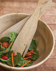 The Be Home Sydney Whale Serving Set features a handcrafted mango wood bowl with spinach, cherry tomatoes, and carrot sticks, plus two fish tail-shaped wooden salad servers on a wooden surface.