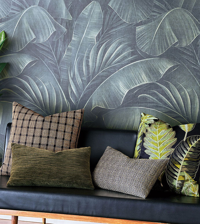 A black bench with four decorative pillows—including Eastern Accents’ BEACHCOMBER CHECK—featuring green, brown, plaid, and textured designs sits before a wall adorned with a bold leafy tropical print.