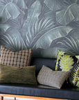 A black bench with four decorative pillows—including Eastern Accents’ BEACHCOMBER CHECK—featuring green, brown, plaid, and textured designs sits before a wall adorned with a bold leafy tropical print.