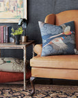 A tan leather armchair with a Barrett Aves Printed pillow by Eastern Accents sits by a rustic side table with books, a plant, and a lamp. Patterned floor cushions and another Barrett Aves Printed pillow complete the cozy scene.