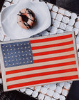 An artistic interpretation of the American flag on a Ben's Garden 5.5" x 8.5" tray, placed on a mosaic tiled surface in a Scottsdale, Arizona bungalow next to a white hat with a floral design.