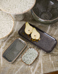 A marble surface displays handmade ceramic plates, bowls, and artisanal serving dishes in neutral tones; the Accent Decor Starling Tray holds two halved pears with seeds and pistachios.