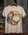 The Stevie Nicks Tee by Made Worn is a cream vintage unisex shirt with a colorful graphic of a long-haired woman, decorative designs, and "Stevie Nicks Live in Concert" text, shown on a wooden hanger before gray lockers.