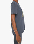 A person wearing the Rowdy, a navy blue 100% cotton crewneck tee by Mother, paired with dark jeans and gold wrist and finger jewelry, shown from the side against a plain white background.