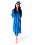 A smiling person, barefoot, wears the Faire Surf Stripe Shirt Dress—blue cotton with buttons—paired with a green-and-white "SURF" cap. They stand hands-in-pockets against a white background.