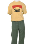 A mannequin models the SURF Tshirt by Hype Factory Inc.—a beige, vintage-washed, 100% cotton tee with a red graphic and "Surf" in black. Features unisex sizing for comfort, paired here with high-waisted green pants.