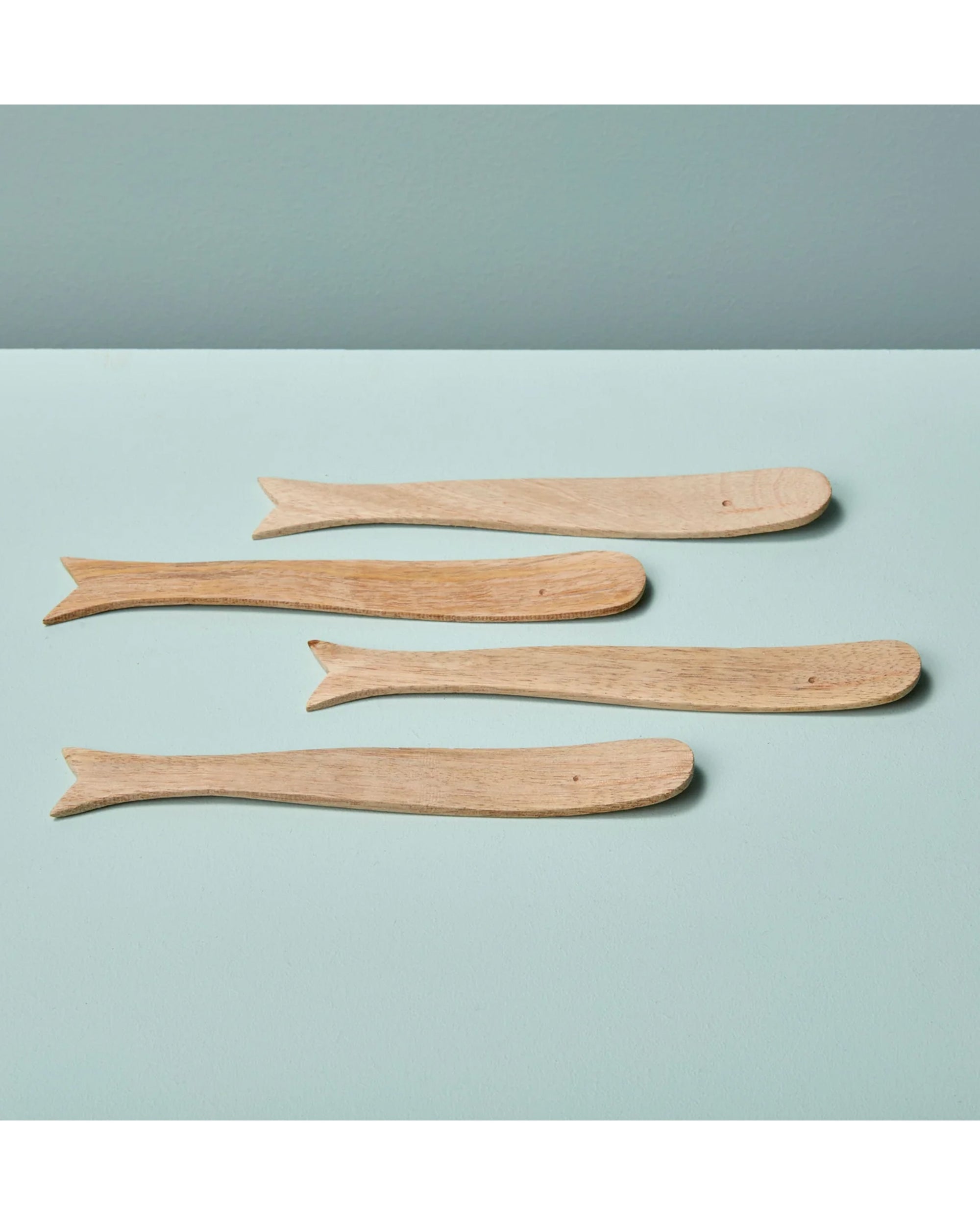 The Be Home 77-102 Sydney Whale Spreaders S/4, handcrafted from sustainably sourced mango wood and shaped like whales, are arranged on a light blue surface against a pale blue background.