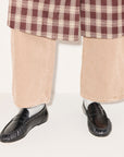 A person stands against a plain light background wearing beige wide-legged pants, a red and white plaid coat, gray socks, and black Sydney Crinkle Loafers by Freda Salvador.