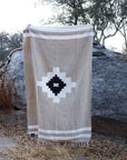The TT Handwoven Blanket Taos Two by Faire, featuring a beige, white, and black geometric diamond pattern, is draped over a large gray rock outdoors among dry grass and trees.