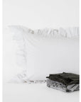 The Matteo Tat Cotton Sham in white, featuring a delicate ruffle border, rests against a white background. In front, gray and black textiles are neatly folded.