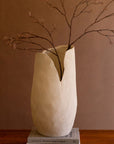 The Bloom Flower Vase L by Kalalou, Inc features a textured cream finish and uneven rim, holding budding branches. This tall white vase sits on stacked books on a wooden surface with a plain brown background.