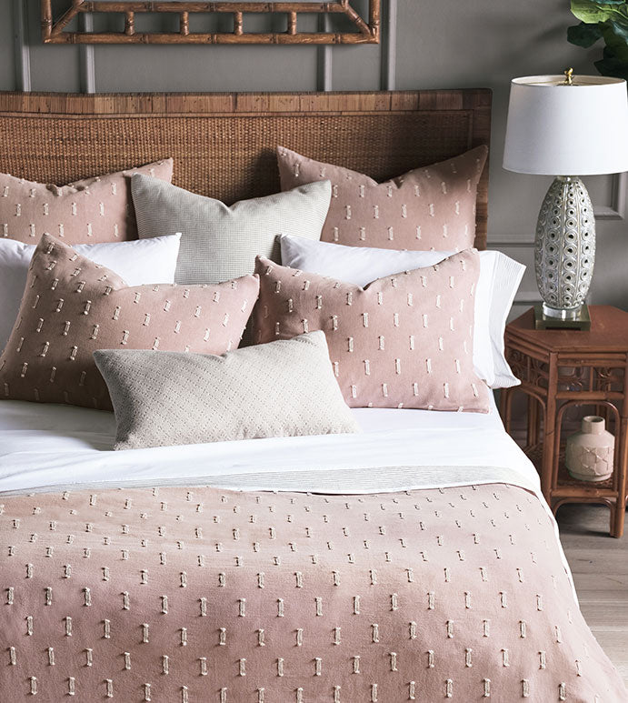 Elegant Bungalow-style bedroom featuring a bed with Eastern Accents Bluff Euro 27x27" bedding in pink and beige, several pillows, a rattan headboard, and a bedside table with a lamp and decor. Subtle gray walls provide a calm backdrop.