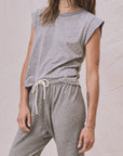 A person stands confidently in The Peak Shoulder Tee by The Great Inc., a sleeveless gray top paired with matching drawstring sweatpants, showcasing tattooed arms against a neutral fabric backdrop, embodying the craftsmanship of U.S.A.-made garments.