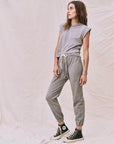 Dressed in a gray jumpsuit with white drawstrings and black high-top sneakers, a person stands against a textured light backdrop. Their relaxed posture enhances their long hair, while The Great Inc.'s handcrafted Peak Shoulder Tee adds a stylish accent from underneath.