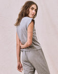 A person with long brown hair stands sideways, looking over their shoulder, wearing The Great Inc.'s stylish sleeveless "The Peak Shoulder Tee" and matching pants against a light fabric backdrop.