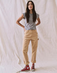 A woman with long dark hair wears The Peak Shoulder Tee by The Great Inc., paired with beige rolled-up pants and red high-top sneakers. She stands with her hands on her hips against a textured beige backdrop.