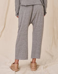 A person wearing The Great Inc.'s The Jersey Crop with gray, relaxed-fit pants featuring a dropped rise and a matching top stands with their back to the camera. They're also wearing tan slip-on shoes against a beige draped fabric background.