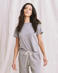 A woman with long dark hair wears The Little Tee by The Great Inc. in grey along with matching drawstring pants, standing before a cream-colored fabric backdrop.