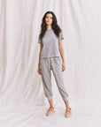 A woman with long dark hair stands before a draped backdrop wearing The Little Tee by The Great Inc. in grey, styled with matching jogger pants and beige moccasin-style shoes, striking a relaxed pose.