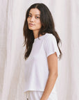 A woman with long dark hair wears The Little Tee by The Great Inc. in white Rancho Stripe, standing before a light fabric backdrop and gazing at the camera with a soft, neutral look.