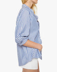 A woman poses in profile against a white background, wearing The Bestie blue and white striped cotton button-up shirt with rolled sleeves from Mother, paired with white shorts.