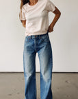 A person stands indoors against a plain wall, wearing Hey Gang’s The Crew 26 light pink short-sleeve t-shirt and vintage fit, high-waisted loose blue jeans with hands on hips; their head is partially out of frame.