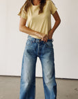 A person indoors fastens the waistband of blue vintage-fit jeans while wearing Hey Gang's The Crew 26, a yellow 100% cotton t-shirt. Their face is out of frame against a plain light-colored background.