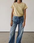 A person stands against a plain light-colored wall, wearing Hey Gang’s The Crew 26 yellow 100% cotton t-shirt and blue high-waisted vintage-fit jeans. Their head is not visible in the image.