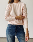A person wearing Hey Gang’s The Sweatshirt 26 in light pink tri-blend fleece and blue jeans stands against a white background, adjusting their sleeve. Their face is not shown.